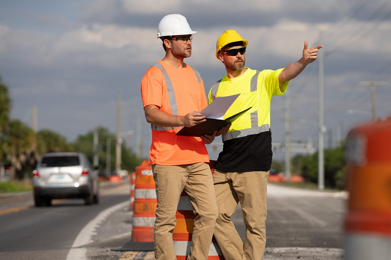 Two road workers review plans beside traffic barrels, with one pointing down the road.